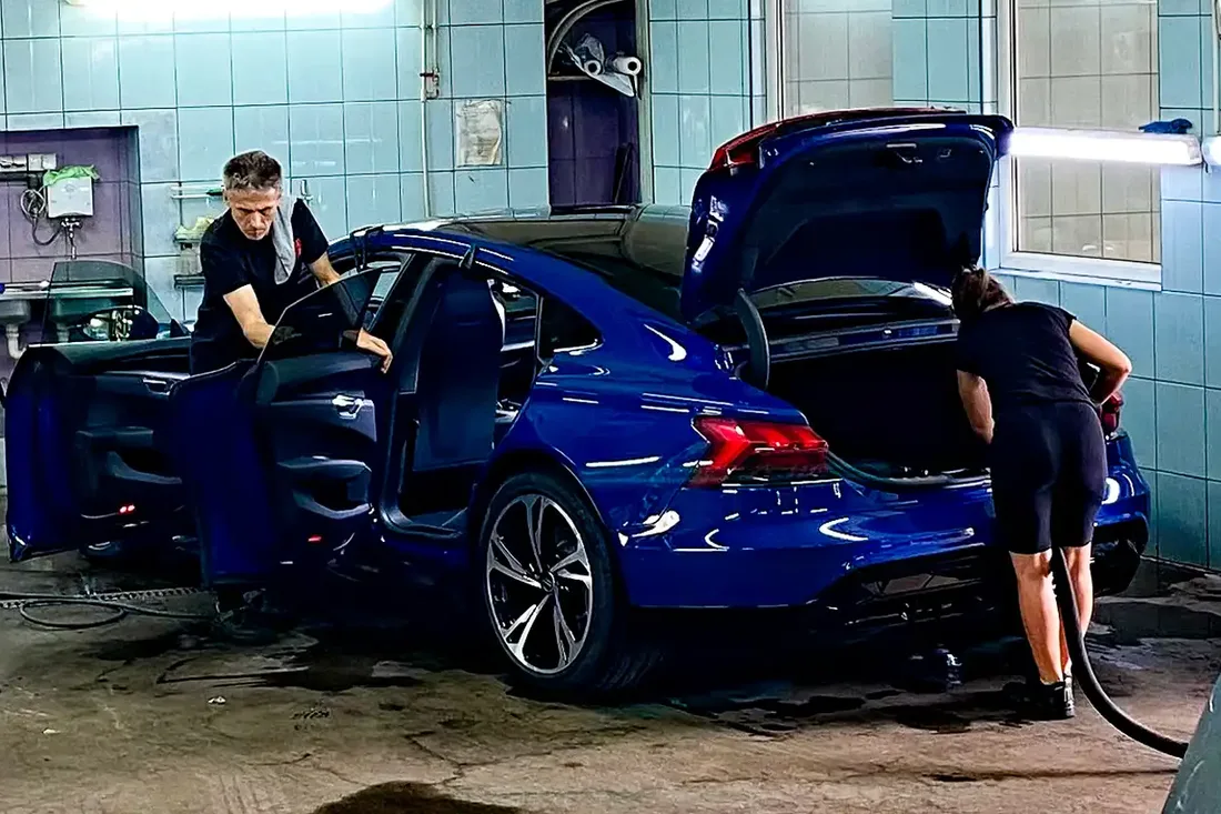 Dwie osoby czyszczą niebieski samochód z otwartymi drzwiami i bagażnikiem.