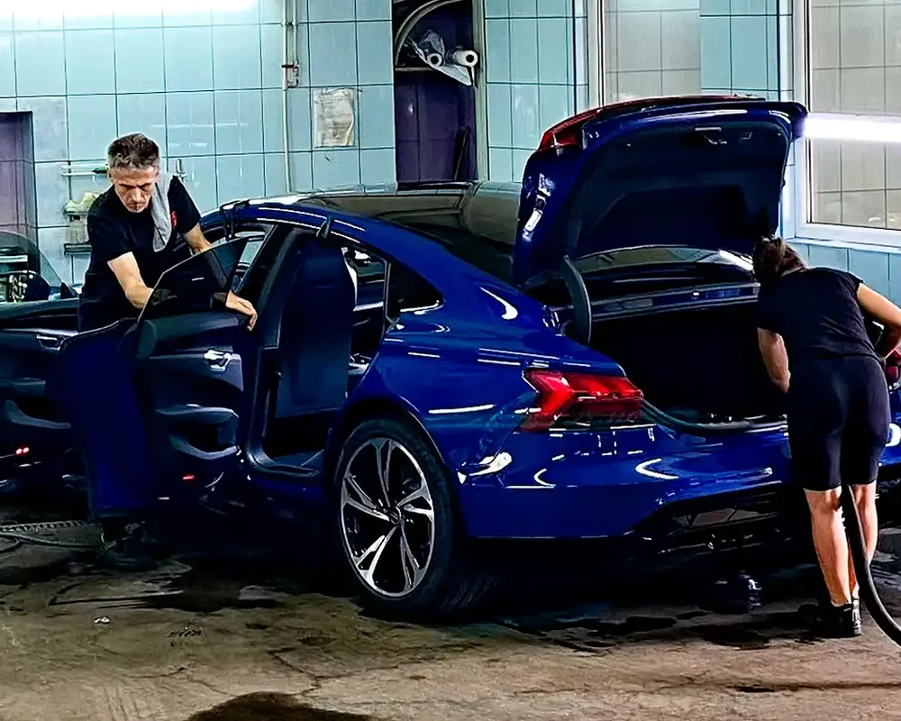 Dwie osoby czyszczą niebieski samochód z otwartymi drzwiami i bagażnikiem.